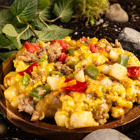 Thumbnail for A wood bowl with a helping of scrambled eggs, vegetables, meat, and cheese. Dirt, rocks, and leaves in the background.