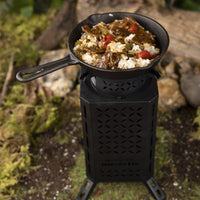 Thumbnail for A cast iron pan with rice, meat, and vegetables cooking on top of the Inferno Pro stove, sitting in a forest of dirt and grass.