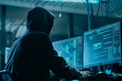 A person wearing a dark hoodie in a low-lit room with a set of computer screens in front of them.