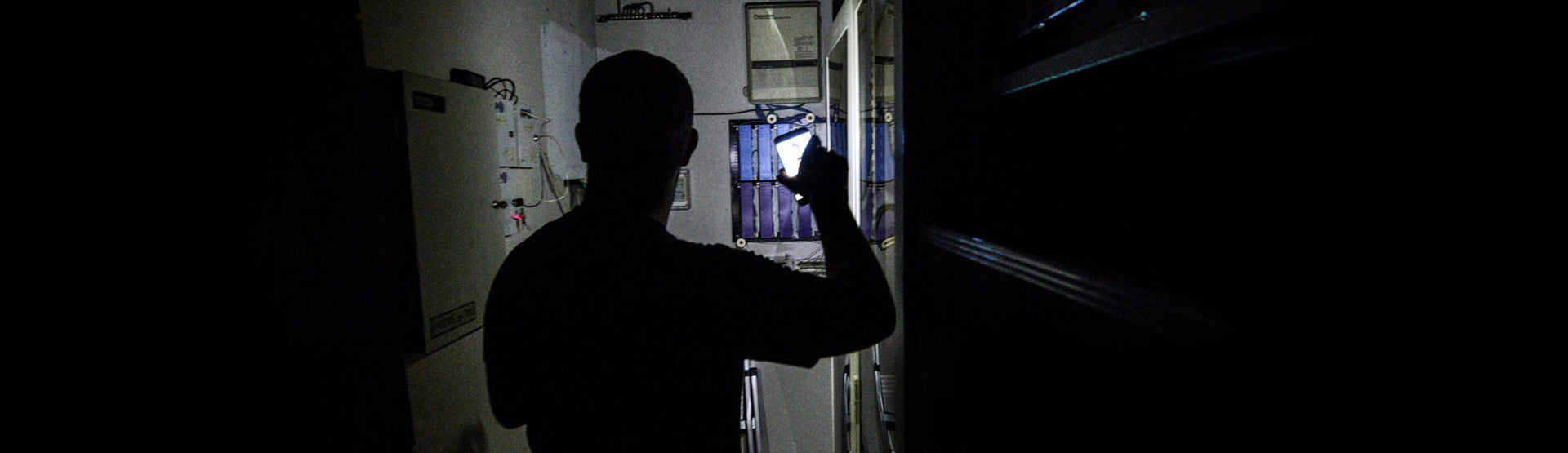 A man navigating a dark hallway with phone flashlight, looking for the fuse box. 